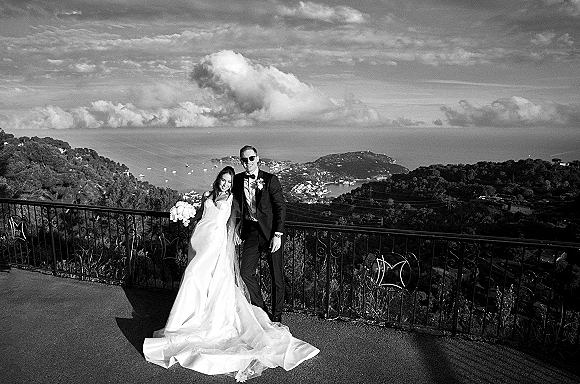 Couple portrait in a black and white wedding portrait style, bride with train and bouquet beside groom in tuxedo and sunglasses at ocean overlook