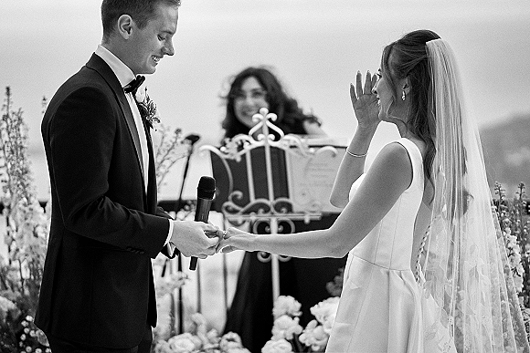 Wedding vows as bride in veil wipes tears while groom in tuxedo holds microphone, hands joined at a mountain outdoor ceremony