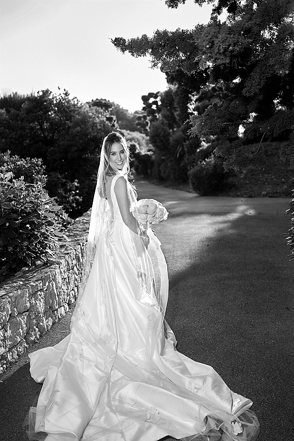 Bridal portrait of a bride looking back, holding a bouquet with a long train and cathedral veil on a garden path by a stone wall