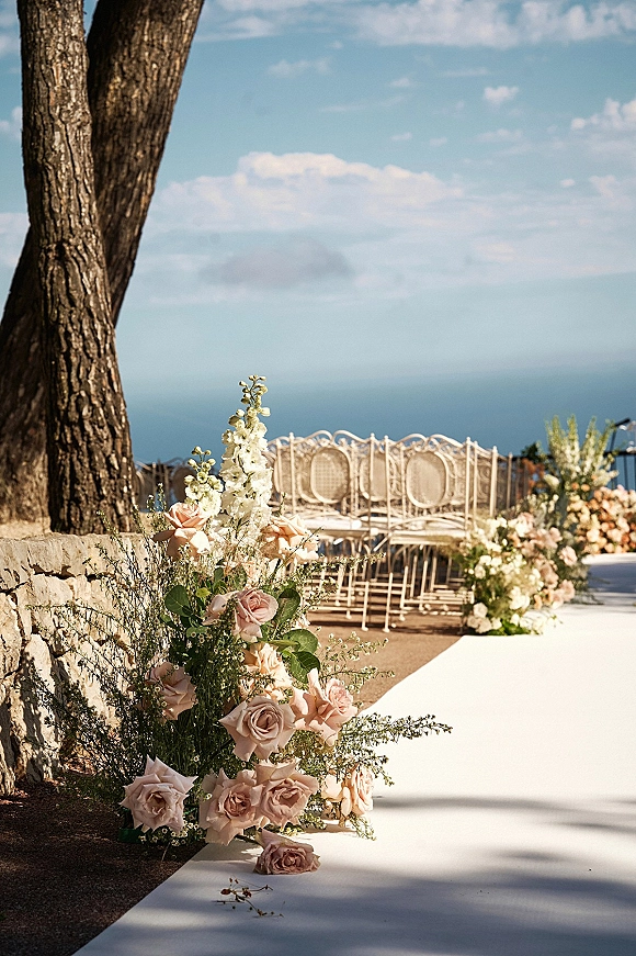 Ceremony aisle decor with wedding aisle flowers of blush roses, white blooms, and greenery lining a white runner on an ocean-view terrace