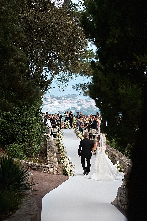 Wedding processional with bride walking down aisle on a white aisle runner, long veil and gown train, groom waiting by stone walls and trees
