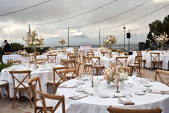Reception tablescape with round tables in white linen, gold pedestal vases, taper candles and floral centerpieces on a mountain-view terrace