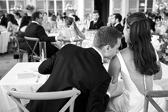 Wedding reception moment as groom kisses bride’s shoulder at their table, candle centerpiece glowing beside linens and guests at banquet tables