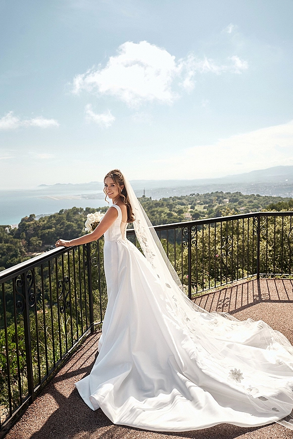 Bridal portrait of a bride holding bouquet on a balcony, long veil trailing as she looks back toward the ocean and hills under clouds