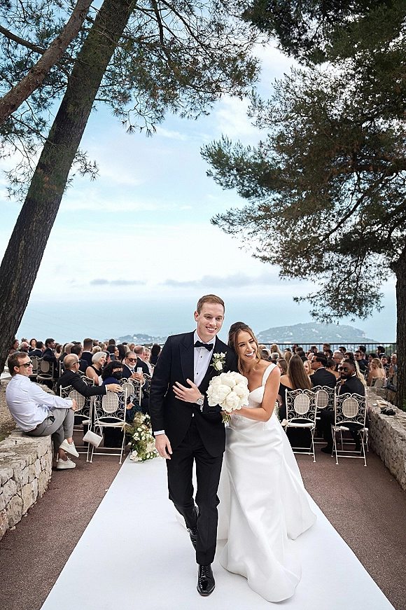 Wedding recessional as bride and groom walk the aisle, bride with white bouquet beside tuxedo groom on an outdoor terrace with ocean view