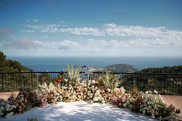 Wedding ceremony backdrop with outdoor ceremony flowers, a white aisle runner, and low florals on a terrace overlooking the ocean view