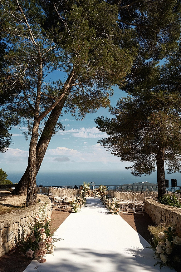 Ceremony aisle decor with outdoor ceremony aisle and white aisle runner, lined with rose and greenery arrangements on an ocean-view terrace