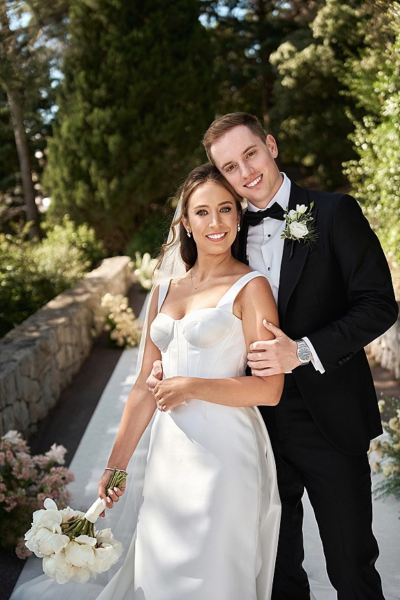 Couple portrait of bride in strapless satin wedding dress and veil holding bouquet as groom hugs her on a garden path by stone wall