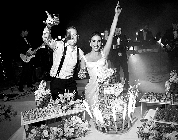Wedding cake cutting as the bride and groom cake table glows with sparklers, dessert display and band stage lighting in a hazy venue