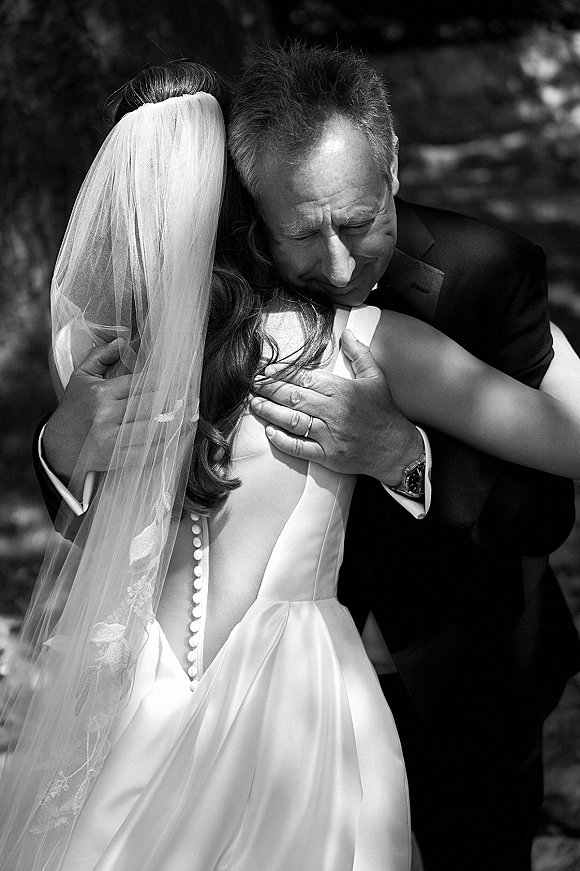 Father daughter wedding hug during a first look, bride in veil and button-back dress embracing dad in a suit amid sunlit trees