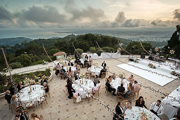 Outdoor wedding reception on a terrace with string lights above round tables, a white dance floor, and stage set against ocean views