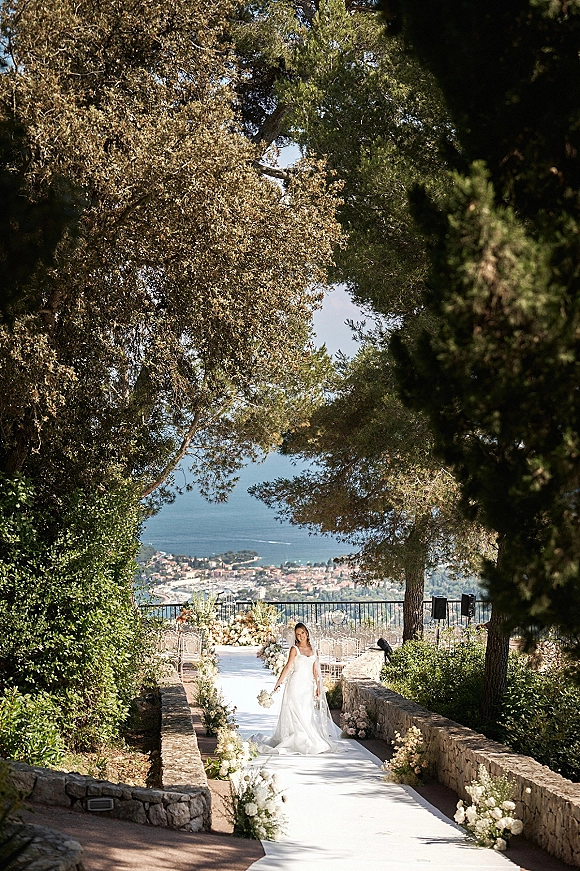 Bridal portrait of a bride walking down the outdoor bridal portrait aisle with veil and bouquet, ocean view beyond stone walls and trees