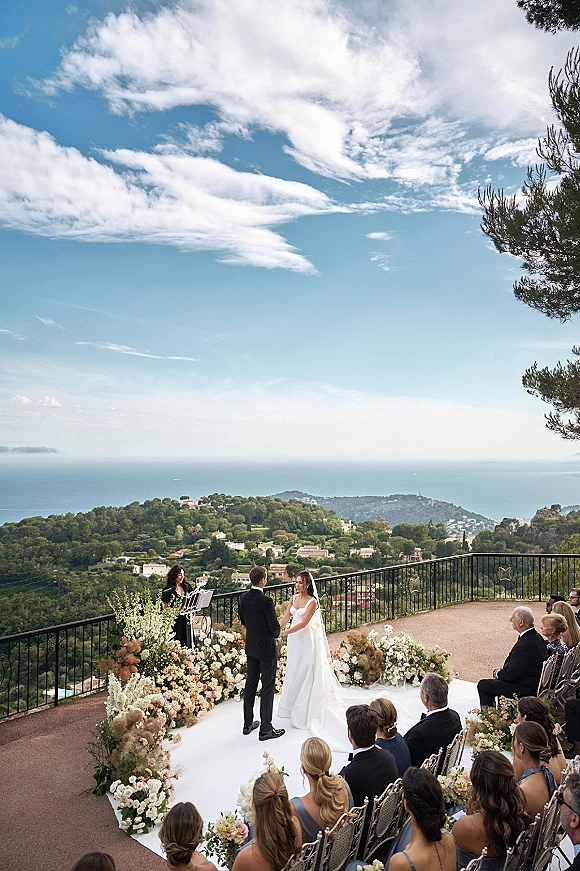 Outdoor wedding ceremony with bride and groom holding hands beneath a floral arch, guests seated on a terrace with ocean view