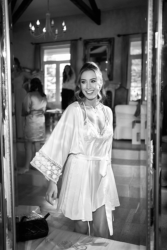 Bride getting ready in a satin bridal robe over lace lingerie, adjusting her necklace by a mirror with window light and chandelier overhead