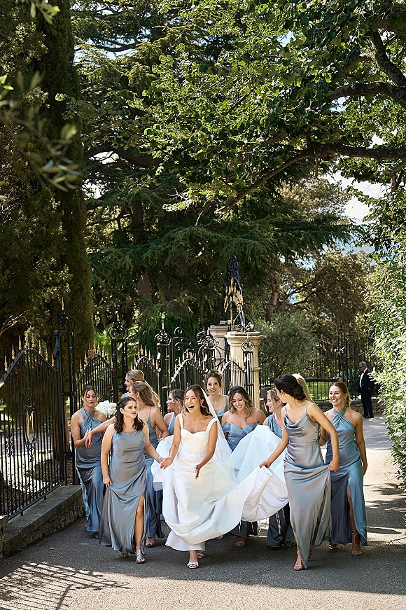 Bride with bridesmaids walking with bride along a sunlit garden path, her veil trailing as they pass wrought iron gates and stone pillars