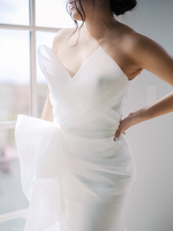 Wedding dress with a strapless neckline and tulle overskirt, sheer wrap bodice detail, styled in soft window light against a white wall