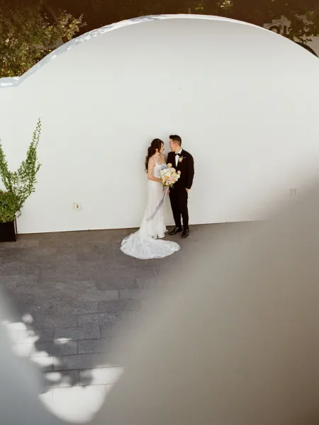 Couple portrait of bride in strapless lace gown holding a pastel bouquet with ribbon tails beside groom in tuxedo against a white curved wall outdoors