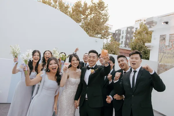 Wedding party photo of bride and groom with wedding party cheering, holding cocktails and bouquets on an outdoor patio by a white stucco wall