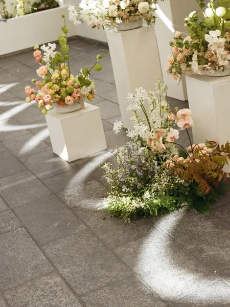 Ceremony floral decor with wedding altar flowers on white pedestal plinths, orchids and greenery on a sunlit stone tile floor with shadows