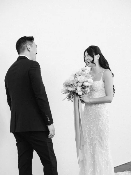 Wedding first look as the groom turns to see the bride in a lace spaghetti-strap gown, laughing and holding a ribboned bouquet against a white wall