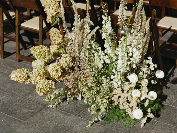 Ceremony aisle florals of white hydrangeas and tall flower spikes with greenery lining wooden chairs on a stone tile floor