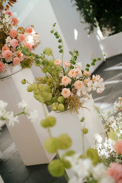 Wedding floral arrangement with peach roses and pink dahlias on pedestal stands in a sunlit outdoor courtyard with white walls and trees