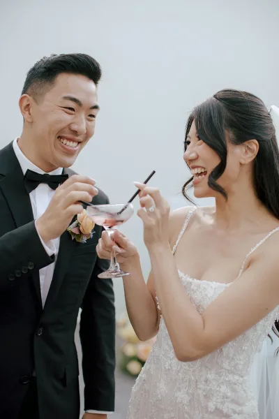 Wedding reception toast as bride and groom clink martini glasses with pink cocktails, bride in veil and groom in tuxedo under open sky