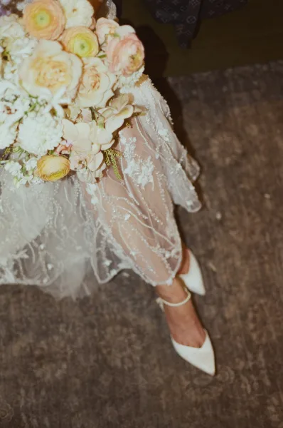 Bridal bouquet of blush and cream roses and ranunculus beside white ankle-strap heels, with sheer lace wedding dress on carpeted floor