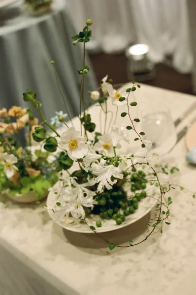 Reception centerpiece with a floral bowl centerpiece of white flowers and greenery in a ceramic bowl, surrounded by candles and glassware on a draped table