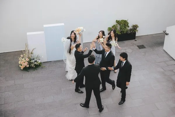 Wedding party photo of bride and bridesmaids with groomsmen raising hands, bride holding bouquet on a stone patio by a white wall