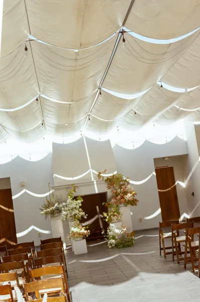 Ceremony setup with indoor wedding ceremony aisle chairs, floral plinths and string lights under a fabric canopy in a sunlit white room