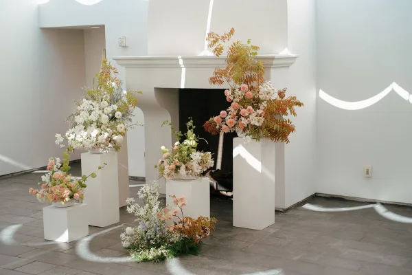 Ceremony altar decor with wedding ceremony backdrop of pedestal florals and greenery framing a fireplace mantel in a bright white room