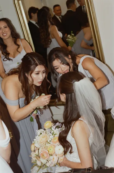 Bride with bridesmaids helping bride adjust her bridal veil, holding bouquets beside a gold-framed mirror in an indoor room