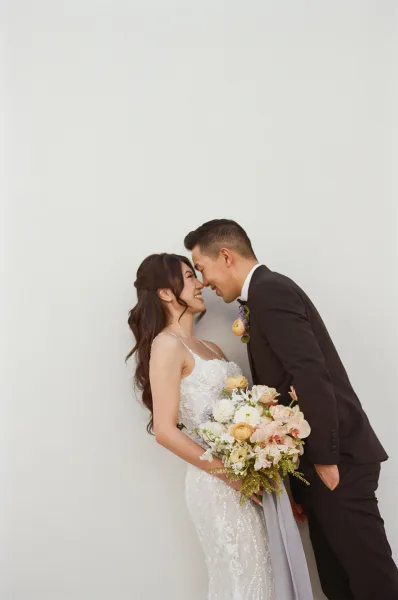 Couple portrait of bride and groom portrait touching foreheads, bride holding a bouquet in lace gown and groom in tux before a white wall