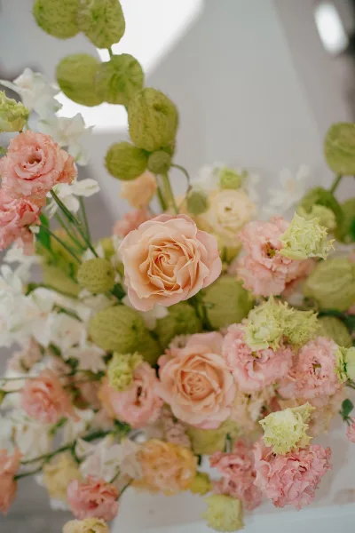 Wedding bouquet of peach rose bouquet blooms with pink carnations, white flowers, green seed pods, and greenery against a white wall in soft light