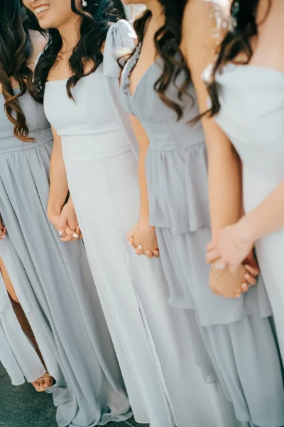 Bridesmaid dresses in dusty blue, mismatched chiffon styles with satin bow strap and ruffle skirts as bridesmaids hold hands on pavement