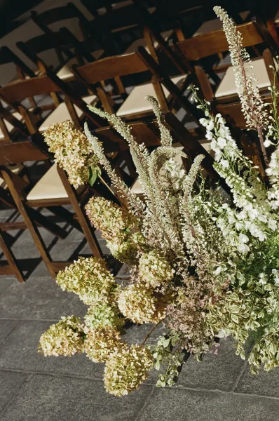 Ceremony aisle flowers with hydrangea blooms and greenery beside wooden folding chairs on a stone floor in an indoor aisle setting