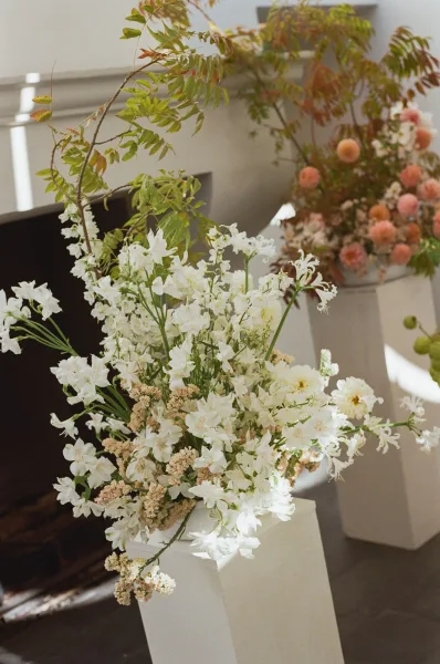 Wedding floral arrangement of white wedding flowers with peach blooms and greenery branches on pedestal plinths in a window-lit indoor venue