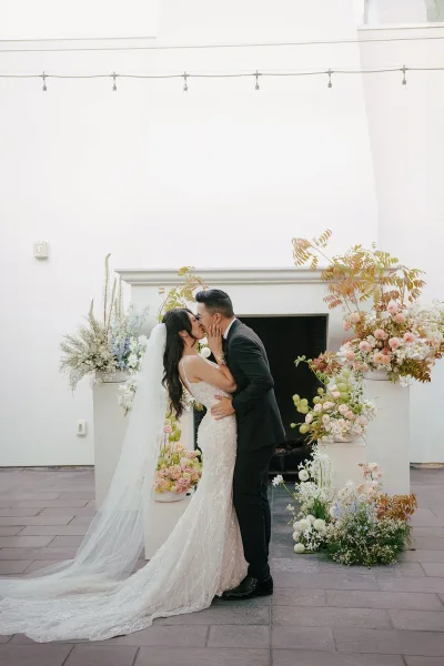 Wedding kiss as bride and groom kissing beneath string lights, her long veil and gown flowing by floral pedestals and fireplace backdrop