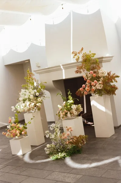 Wedding floral display with ceremony flower pedestals on white plinths, greenery and vases under a skylit indoor courtyard with string lights
