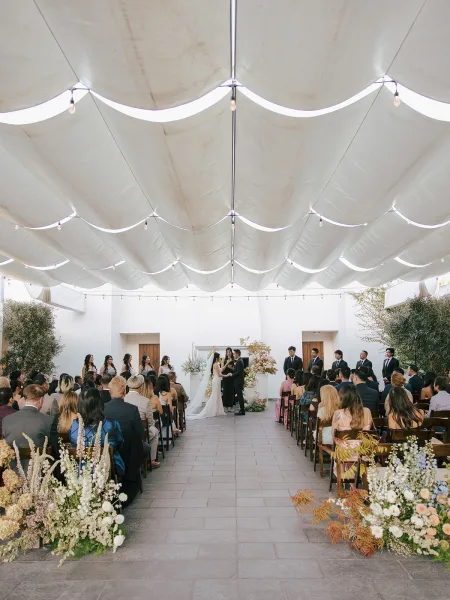 Wedding ceremony with outdoor wedding ceremony aisle lined in blush florals, under draped fabric and string lights in a white courtyard