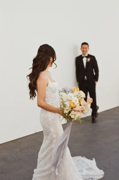 First look moment as bride approaches groom, holding a bouquet with ribbon streamers, lace train trailing by a white wall