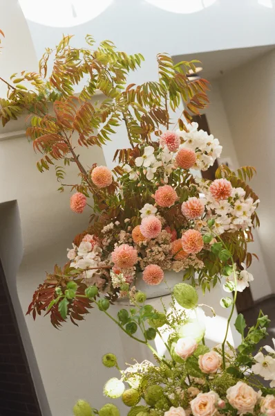 Wedding floral arrangement in a ceramic pedestal bowl with dahlias, roses, white blooms, and greenery in a sunlit interior alcove