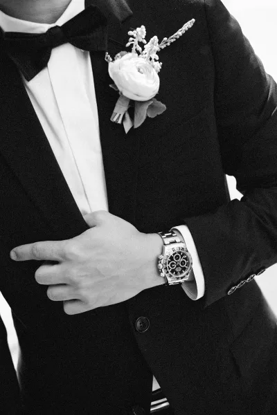 Groom portrait in a black tuxedo with bow tie, white boutonniere on lapel, and wristwatch against a plain light backdrop