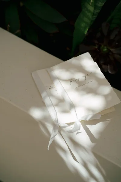 Wedding vows booklets tied with a white ribbon, featuring calligraphy on deckle edge paper on a cream tabletop with greenery and sun shadows