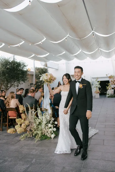 Wedding recessional as bride and groom walk the aisle holding hands, bride lifting a bouquet with ribbon streamers under string lights canopy