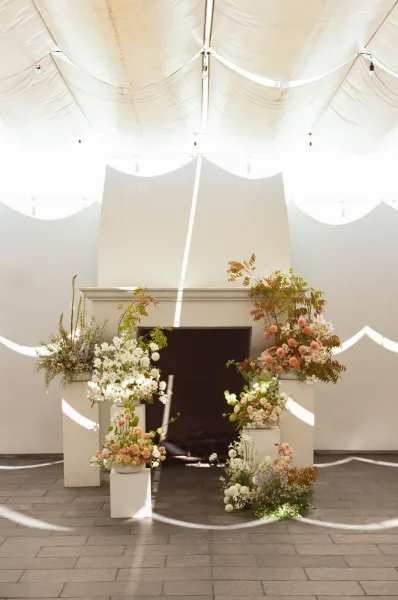 Wedding ceremony backdrop with ceremony altar flowers on pedestal plinths, draped tent ceiling and string lights by a fireplace mantel
