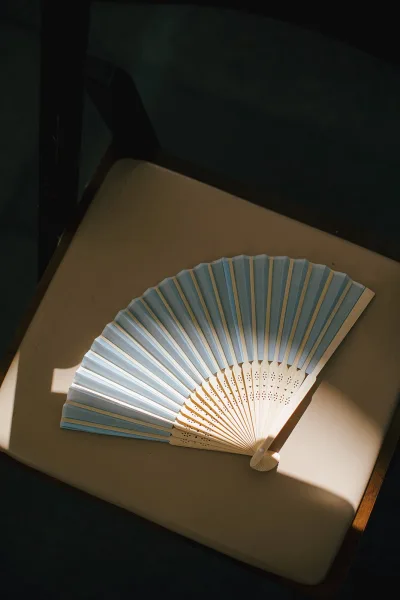 Wedding hand fan, a folding wedding fan in blue and white resting on an upholstered chair with sunlit shadows along the wood frame