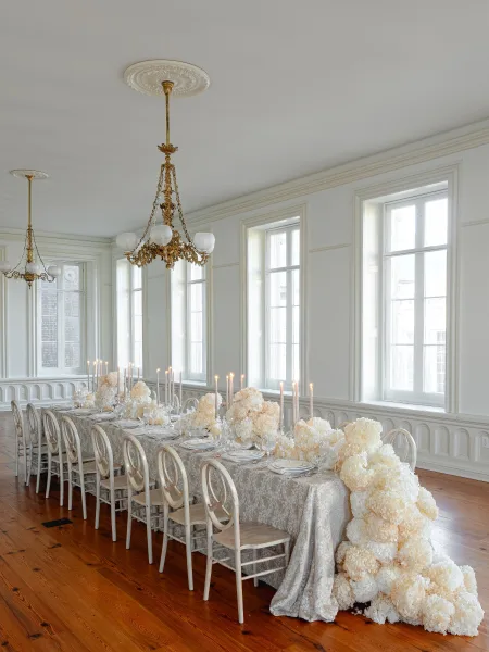 Reception tablescape with a long banquet table setup, ivory floral runner and taper candles beneath chandeliers in a bright white-paneled room