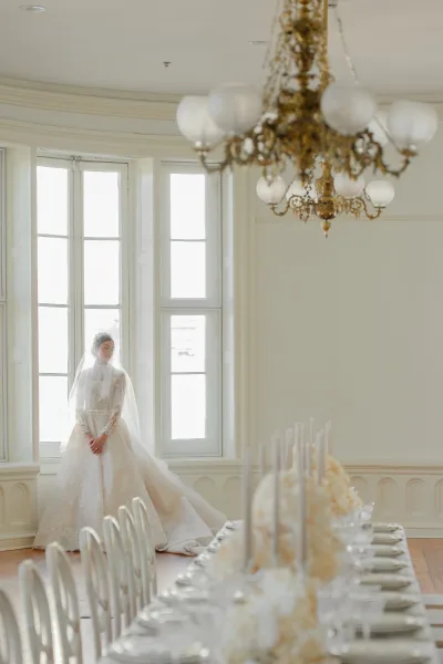 Bridal portrait of a bride in a lace wedding dress with long veil and tiara beside a chandelier-lit banquet table by tall windows